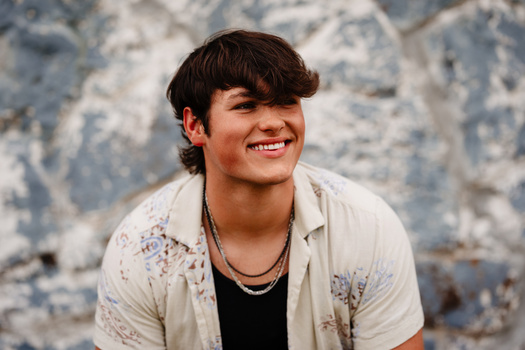 Young man smiling at camera wearing casual button-up shirt over black tee with beaded necklace.