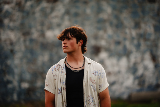 Young man in patterned shirt looking to the side against textured blue and gray wall.