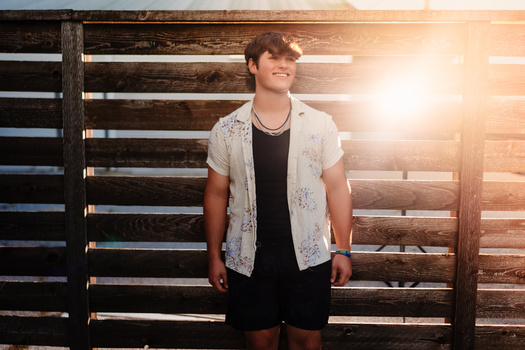 Young man in casual layered clothing standing against backlit wooden fence slats.