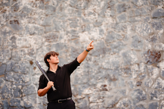 Man in black shirt pointing upward while holding a baseball bat against a weathered stone wall.