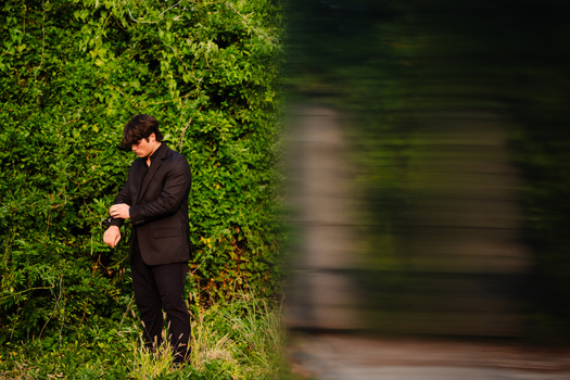 Man in dark blazer standing against dense green ivy wall, holding phone and looking downward.
