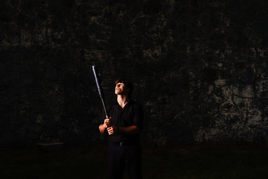 Person in black clothing holding a baseball bat, spotlit against a dark textured wall.
