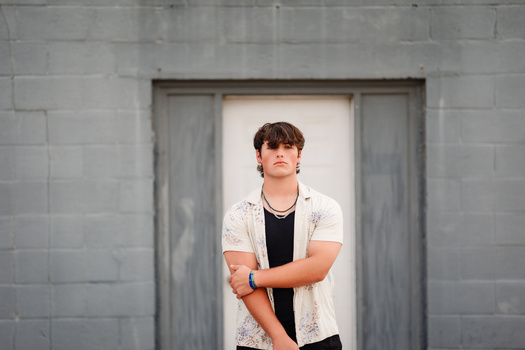 Young man in white patterned shirt standing in doorway against gray concrete wall.