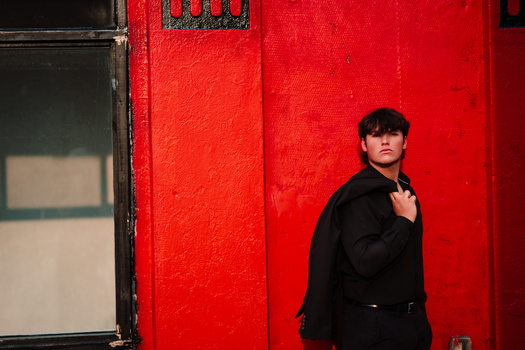 Person in black blazer standing against a vibrant red wall, adjusting their collar.