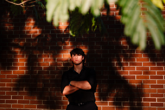 Person in black shirt posing with crossed arms against brick wall with warm lighting and foliage overhead.