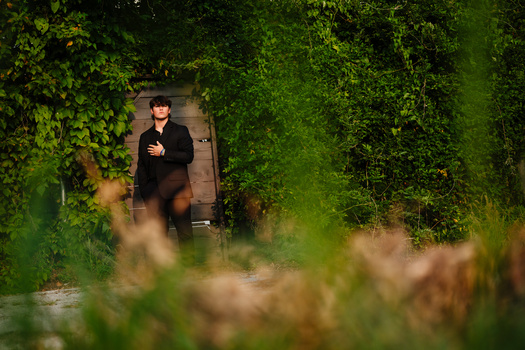 Man in dark clothing standing in doorway framed by lush green ivy and overgrown vegetation.