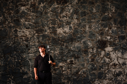 Man in black clothing holding a baseball bat against a weathered concrete wall.
