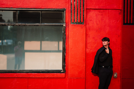 Woman in dark blazer standing against vibrant red wall with large window beside her.