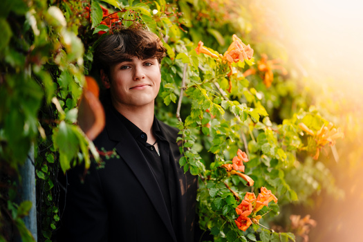 Young man in black jacket posed among flowering vines with warm golden sunlight filtering through foliage.