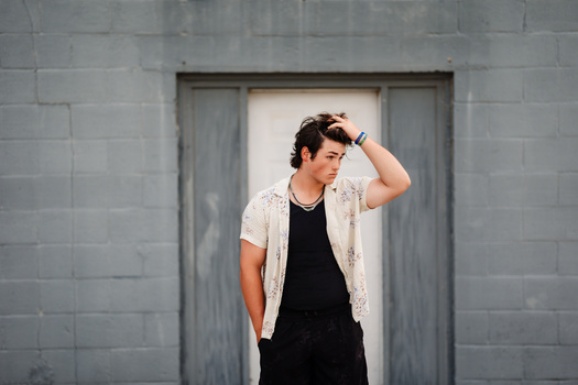 Man in black shirt and cream cardigan posing against gray brick wall with hand to hair.