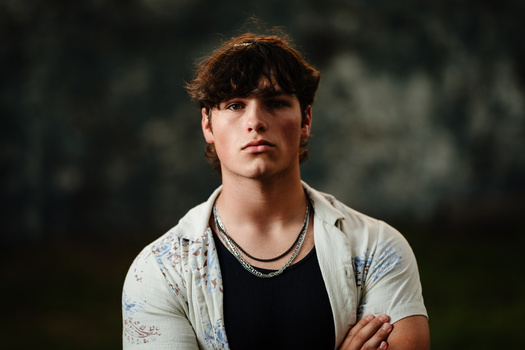 Young man with dark hair and bangs wearing a light button-up shirt over a black t-shirt, posed outdoors with arms crossed.