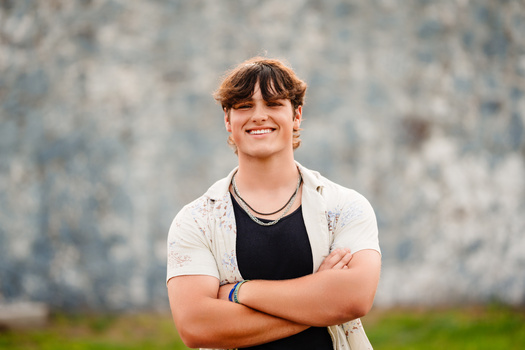 Young person with reddish-brown hair smiling at camera with arms crossed, wearing black shirt and light cardigan against textured gray wall.