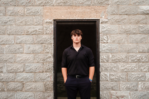 Young man in black V-neck shirt standing in dark doorway of stone brick wall.