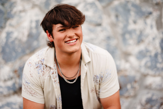 Young man smiling at camera wearing casual layered shirt over black tee against textured wall.