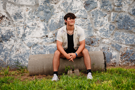 Man sitting on a concrete pipe against a weathered stone wall.