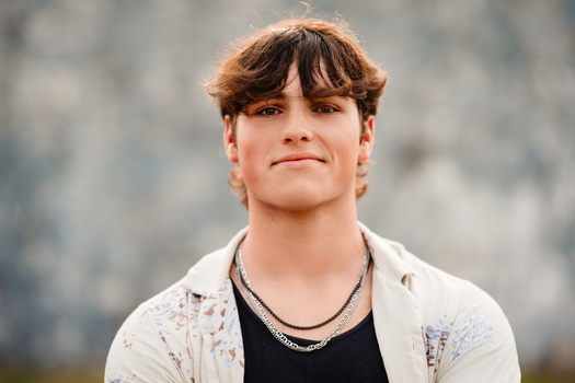 Young man with reddish-brown hair and bangs wearing a light patterned shirt over a black t-shirt and chain necklace.