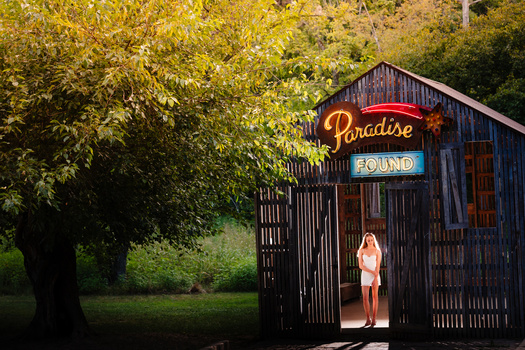 Woman in white dress standing in doorway of dark wooden structure with neon Paradise and Found signs, surrounded by golden autumn foliage.