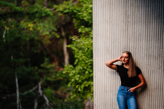 Woman in black shirt and jeans posing against corrugated metal wall with green foliage behind.
