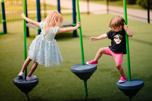 Two young children playing on green playground equipment with suspended balance platforms on artificial turf.