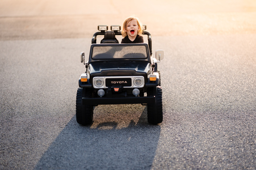 Child in toy Toyota Jeep driving on desert road with excited expression.
