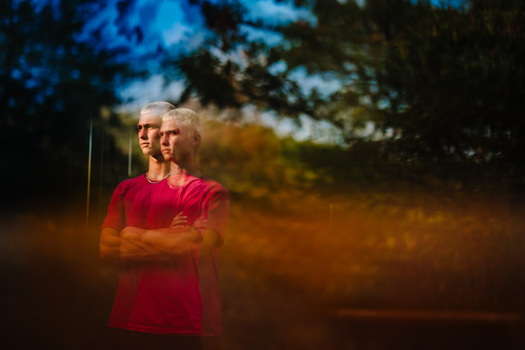 Person in red shirt with white hair wearing sunglasses, standing with arms crossed against blurred natural background.