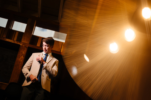 Man in blazer and tie speaking at microphone under warm stage lights.