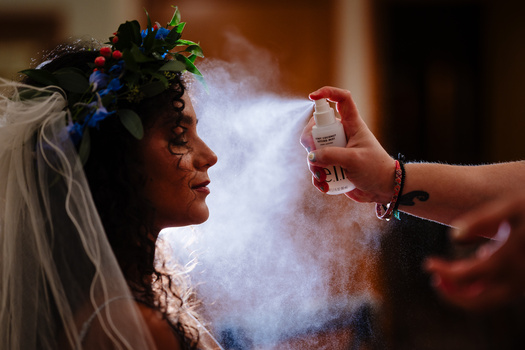 Bride in profile wearing floral headpiece and veil while makeup artist applies setting spray to her face.