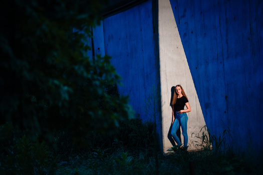 Woman in black top and jeans standing against a white vertical stripe on a blue textured wall.
