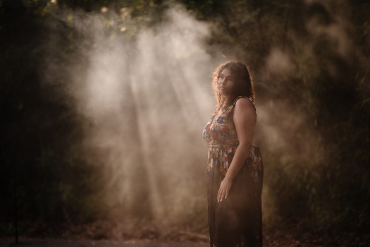 Woman in patterned dress standing in forest with dramatic volumetric light rays filtering through trees.