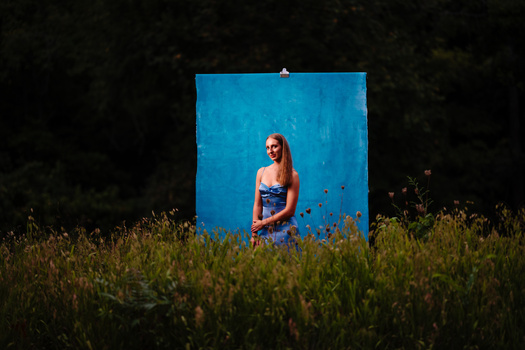 Woman in sunglasses and blue top standing against a vibrant blue backdrop in an overgrown field.