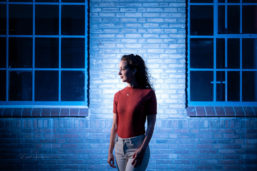Woman in red shirt standing against blue brick wall with grid windows behind her.