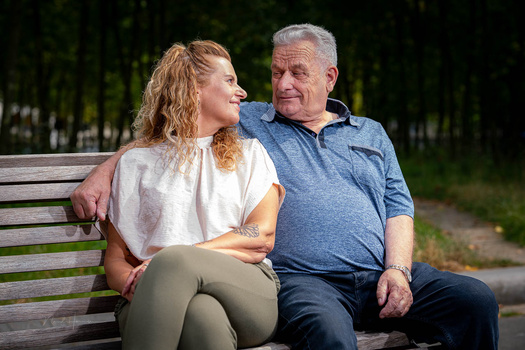 Older man and younger woman sitting together on a wooden bench outdoors, looking at each other affectionately.