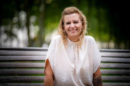 Woman with blonde wavy hair smiling at camera while sitting on a bench outdoors.