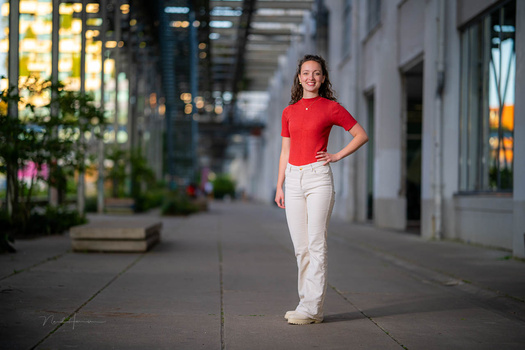 Woman in red shirt and white pants standing on urban sidewalk at dusk.