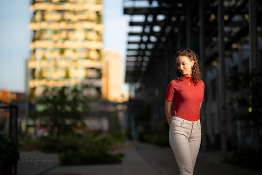 Young woman in red shirt and beige pants standing in urban alley with golden-hour sunlight.