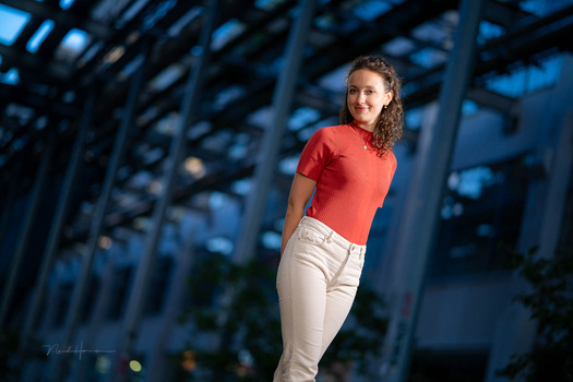 Woman in red shirt and cream pants posing against modern glass building at dusk.