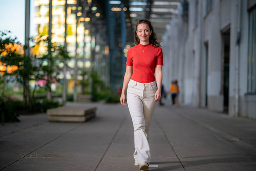 Woman in red shirt and white pants walking toward camera on urban sidewalk.