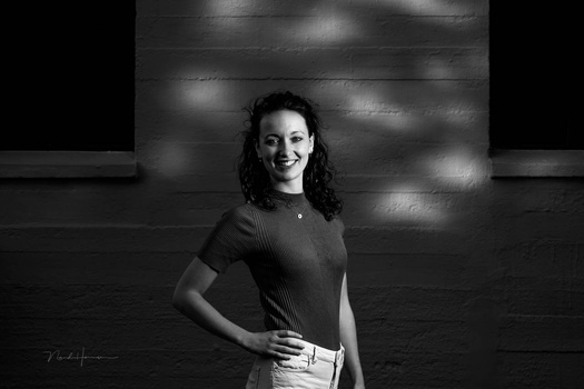 Woman with curly dark hair smiling at camera against textured brick wall with dramatic side lighting.