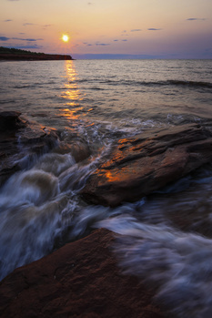 Sunset over ocean with waves washing across dark rocks in foreground.