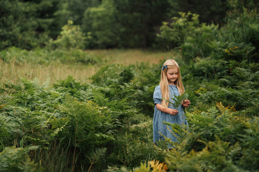 Young girl with blonde hair standing among green plants and vegetation in an overgrown field.