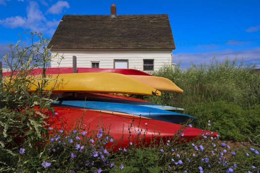 White farmhouse with red trim stacked behind three colorful kayaks and flowering shrubs.