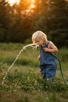 Toddler in denim overalls spraying water from a garden hose at golden hour.