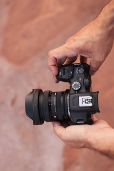 Hands holding a compact thermal imaging camera with attached lens against a neutral background.