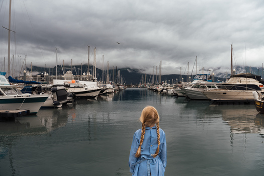 Person with red hair viewed from behind standing at a marina overlooking moored sailboats under overcast sky.