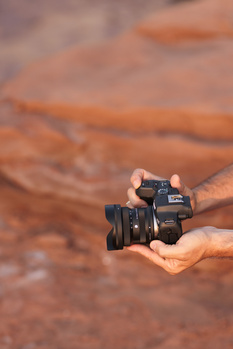 Hand holding a compact video camera with flip-out monitor against blurred earth-toned background.