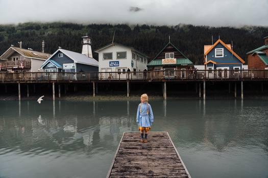 Child in light blue dress standing alone on wooden pier extending into calm gray-green water, with colorful stilt houses and forested hills in misty background.