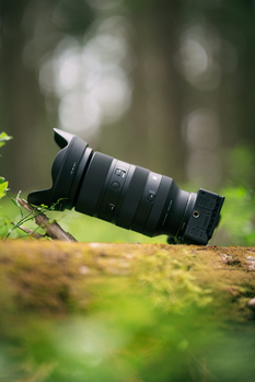 Black telephoto lens positioned on moss-covered ground with blurred green foliage background.