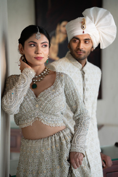Woman in beaded crop top and skirt with man in white turban and traditional outfit posed together indoors.
