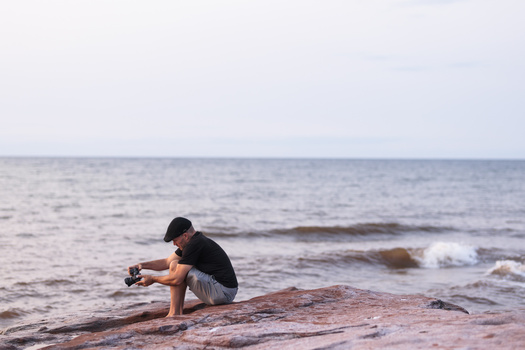 Person crouching on a rocky shoreline, holding a camera while facing the ocean with waves in the background.