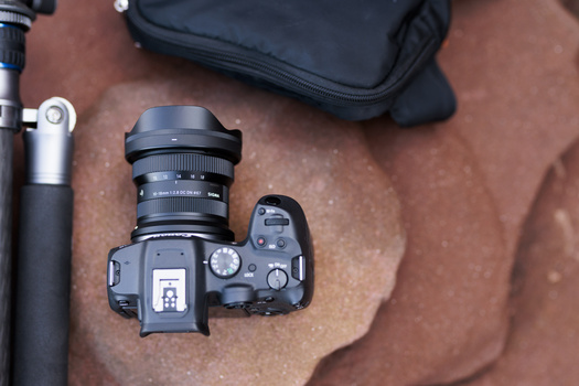 Mirrorless camera with wide-angle lens mounted on brown fabric surface next to tripod and black camera bag.